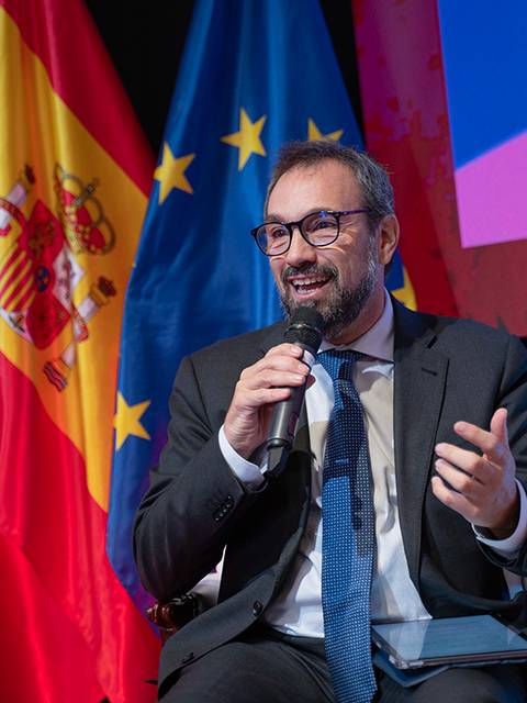 Un hombre con gafas sonríe mientras habla en un evento con banderas de España y la Unión Europea de fondo.