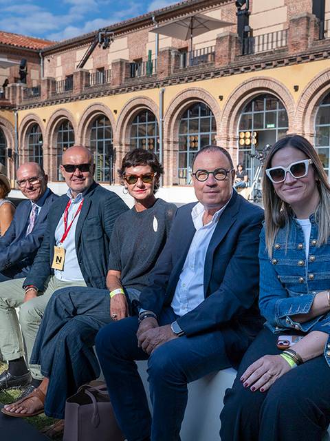 Un grupo de personas sentadas en un evento al aire libre, con un edificio histórico al fondo.