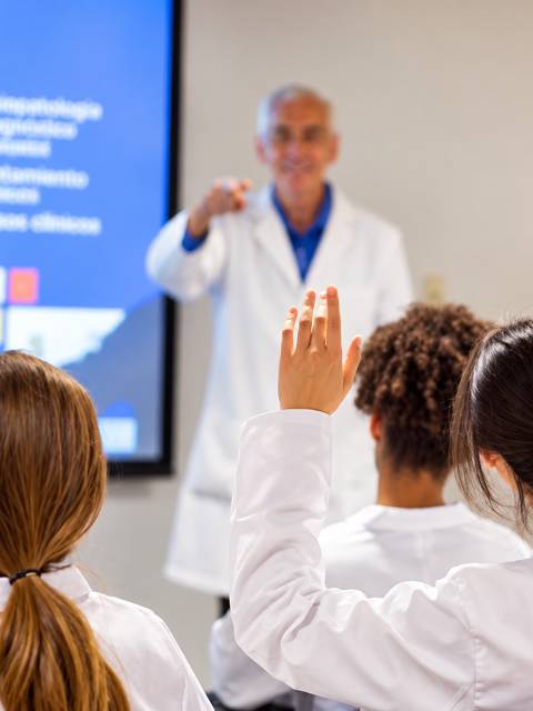 Un grupo de estudiantes en bata blanca levanta la mano durante una clase impartida por un profesor.
