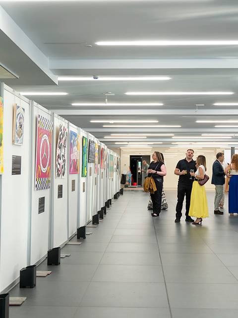 Una exposición de arte con varias obras colgadas en paneles y un grupo de personas conversando.