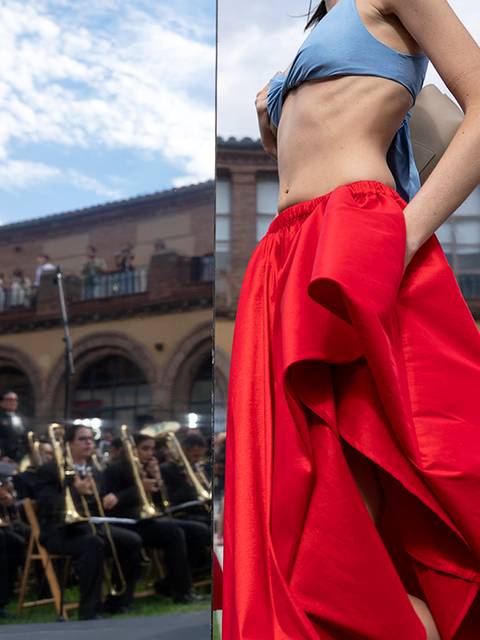 Un modelo presenta un atuendo colorido en un desfile de moda al aire libre, con un fondo de músicos y un público atento.