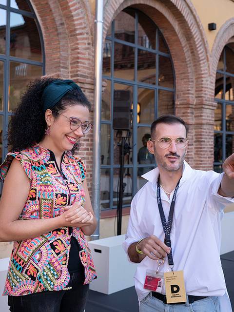 Dos personas conversan en un entorno exterior, con una arquitectura de fondo.