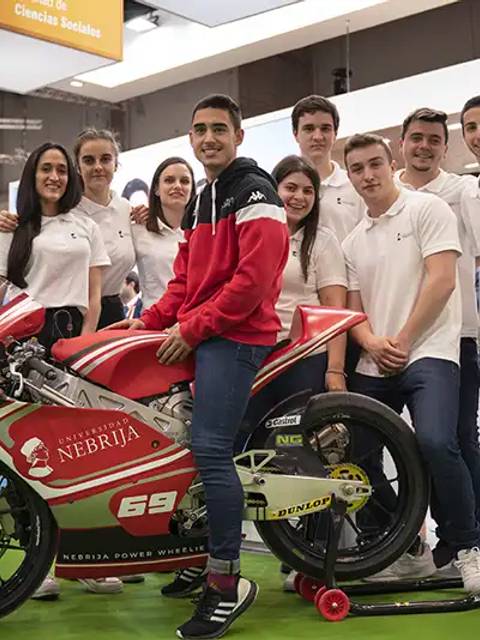 Un grupo de jóvenes sonrientes se reúne alrededor de una moto de competición en un evento.
