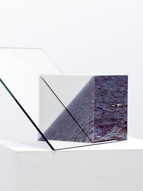 La imagen muestra una escultura geométrica minimalista compuesta por cristales de vidrio y una representación de agua.