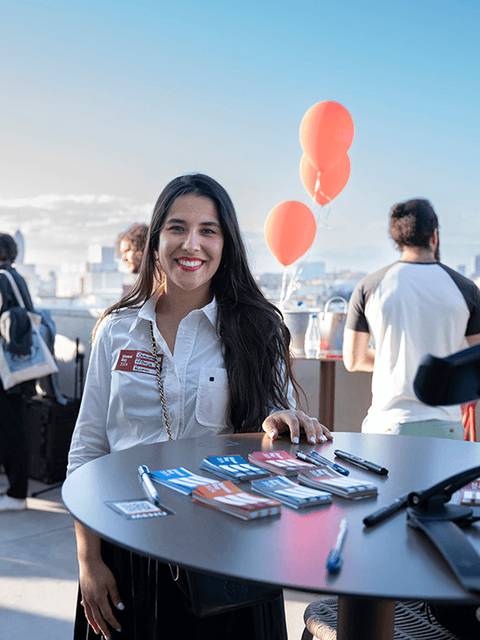 Una mujer sonriente se encuentra en un evento al aire libre, junto a una mesa con materiales promocionales y globos naranjas en el fondo.