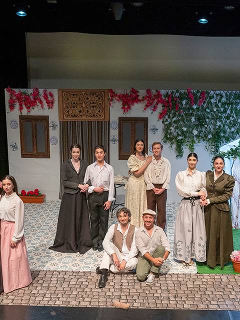Grupo de actores posando en un escenario decorado con elementos naturales y un fondo teatral.