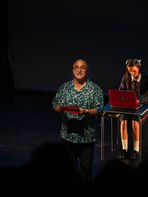 Un hombre presenta en el escenario mientras una niña trabaja en una computadora portátil.