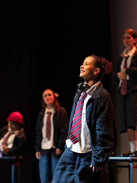 Un grupo de jóvenes actores se presenta en un escenario, vestidos con uniformes escolares, mientras uno de ellos actúa con una expresión de alegría.