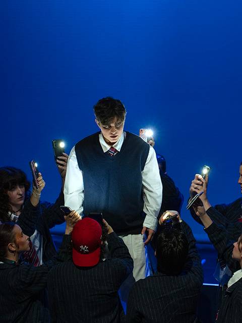 Un grupo de personas rodea a un joven iluminado por luces de teléfono móvil en un escenario oscuro.