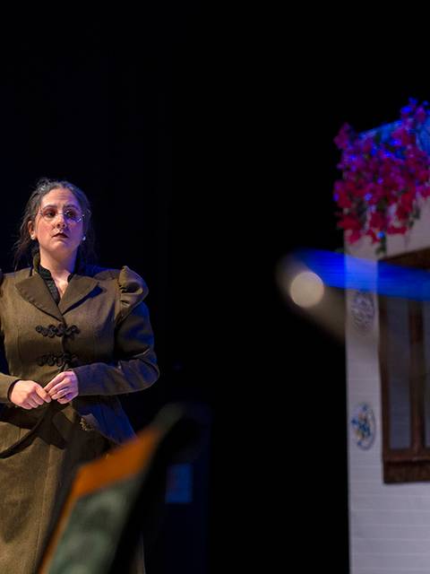 Una mujer vestida con un abrigo oscuro se encuentra de pie en un escenario, con un fondo decorado con flores y elementos de una casa.