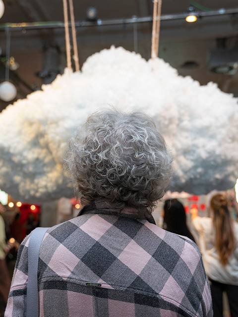 Una mujer mayor observa una nube de algodón suspendida en un ambiente artístico con personas alrededor.