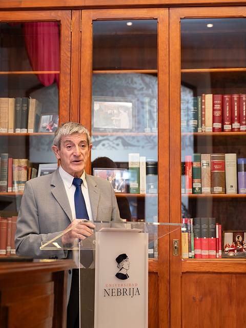 Un hombre habla frente a un atril en una biblioteca con estanterías llenas de libros.