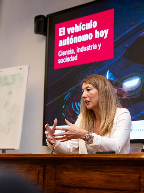 Una mujer habla sobre el vehículo autónomo en una presentación en frente de una pantalla que muestra un automóvil y el título del evento.