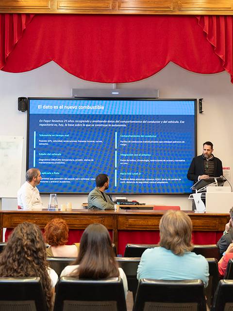 Una presentación en un aula con un ponente frente a una pantalla grande y una audiencia atenta.