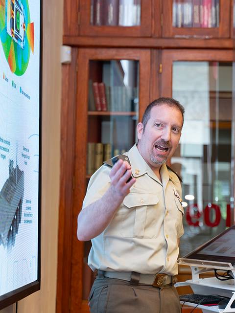 Un hombre con uniforme está dando una presentación frente a una pantalla con gráficos y fotografías.