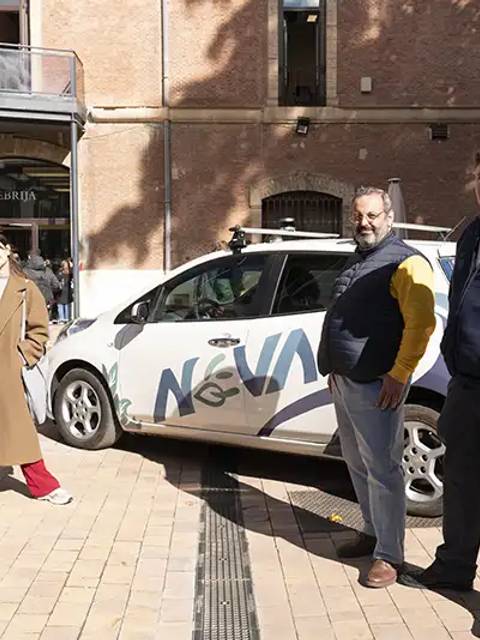 Cuatro personas posan junto a un vehículo en una plaza al aire libre.