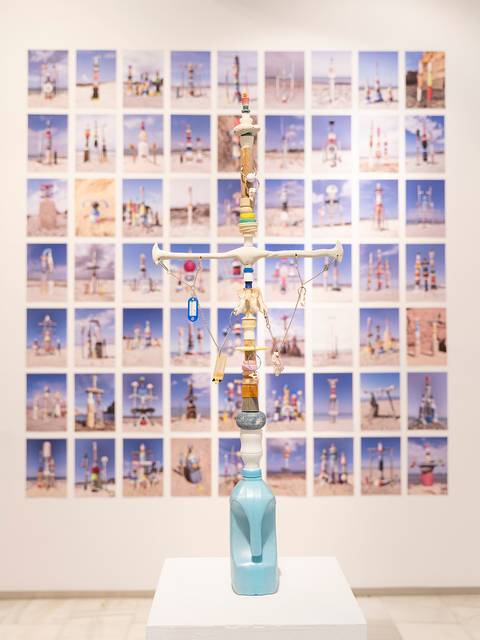 La imagen muestra una escultura realizada con objetos reciclados en una galería de arte, con fotografías de estructuras similares en el fondo.