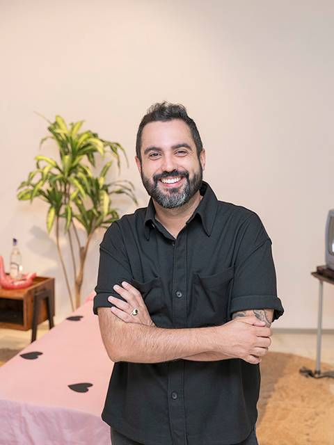 Un hombre sonriente posa frente a una decoración con una mesa y una planta en un ambiente artístico.