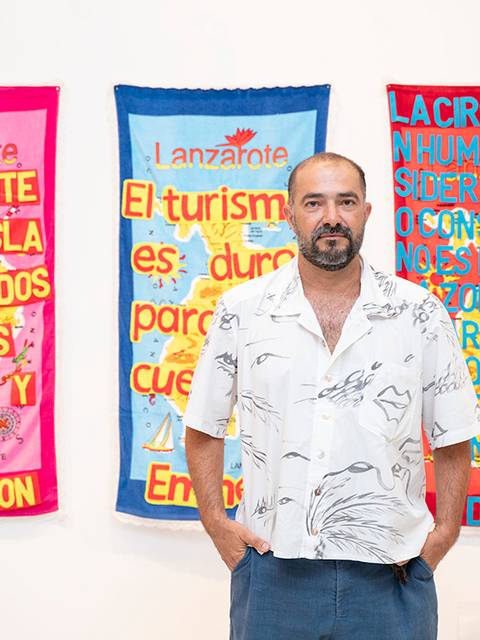 Un hombre de pie frente a tres pancartas coloridas con mensajes sobre turismo y crítica social.