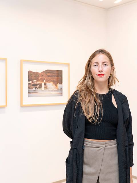 Una mujer con cabello largo y suelto posa en una galería de arte con fotografías en la pared.