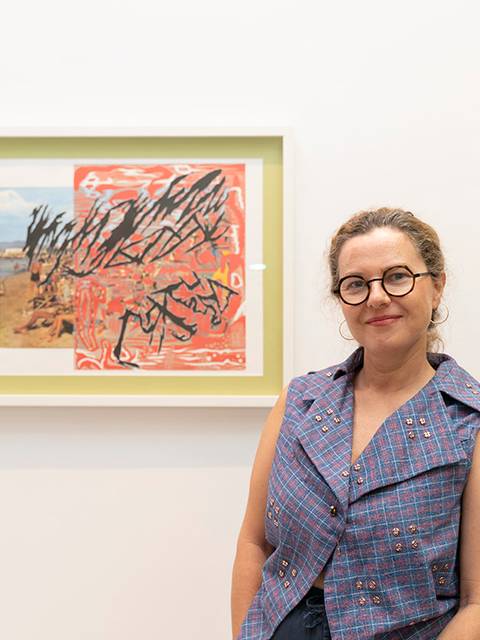Una mujer sonriente posa frente a una obra de arte enmarcada en la pared de una galería.