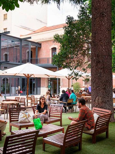 El lugar muestra un jardín con mesas y sillas, donde varias personas están conversando y disfrutando del ambiente al aire libre.