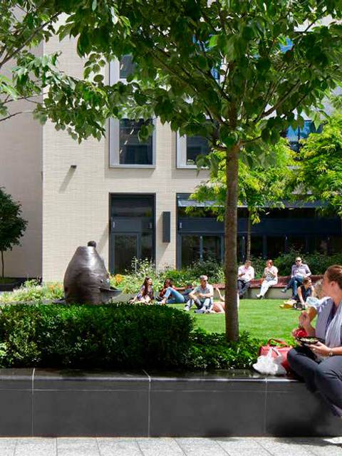 Una mujer sentada en un banco rodeada de personas en un espacio verde con esculturas y prédios modernos.