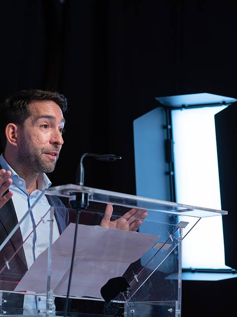 Un hombre está hablando en un evento en el escenario, frente a un micrófono y con una iluminación adecuada.
