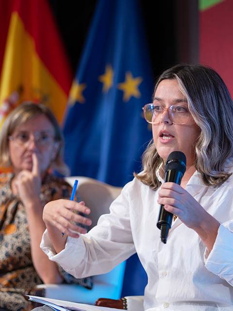 Una mujer habla en un panel mientras otra persona la escucha atentamente en un evento con banderas de España y la Unión Europea de fondo.