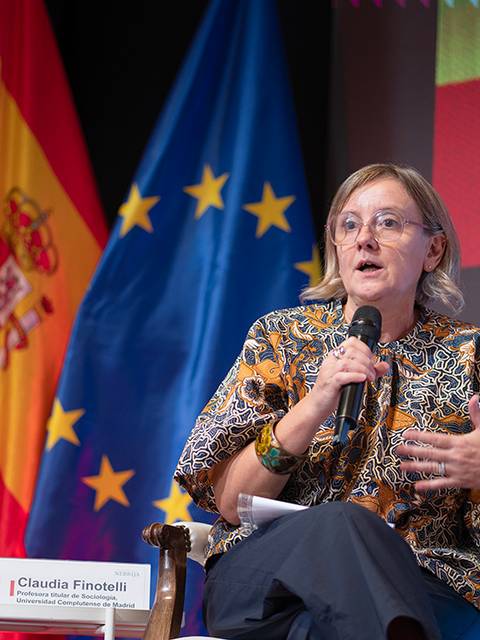 Una mujer habla en un evento mientras se encuentran visibles varias banderas de España y de la Unión Europea.