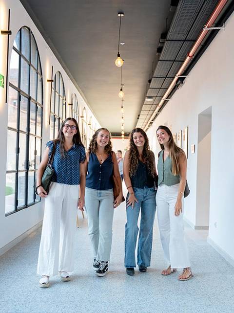 Cuatro mujeres posan sonrientes en un pasillo con paredes blancas y ventanas grandes.