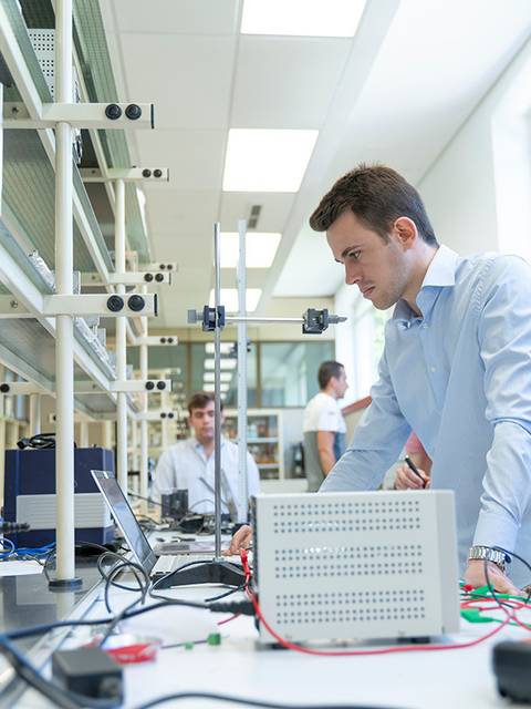 Un estudiante trabaja en un laboratorio con equipos electrónicos y otros compañeros al fondo.