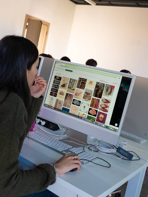 Una mujer joven está buscando imágenes en un ordenador en un aula llena de computadoras.