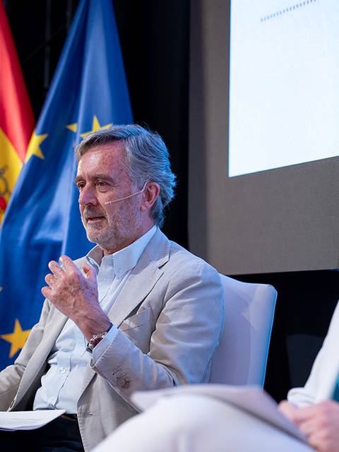 Un hombre de cabello canoso y una mujer con cabello rubio están hablando en un evento con banderas de España y la Unión Europea de fondo.