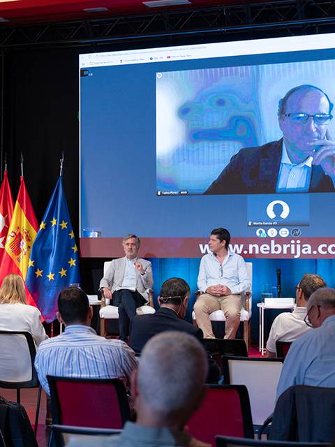 Un panel de discusión con participantes en un escenario acompañado de una videoconferencia proyectada en una pantalla grande.