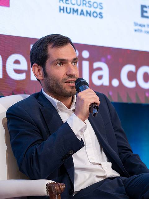 Un hombre sentado en un escenario hablando con un micrófono en una conferencia.