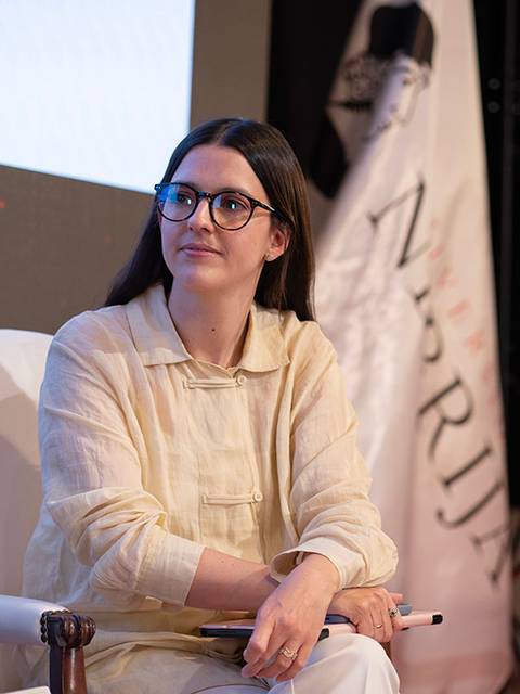 Una mujer sentada en un evento, luciendo una camisa clara y gafas, mientras observa atentamente.