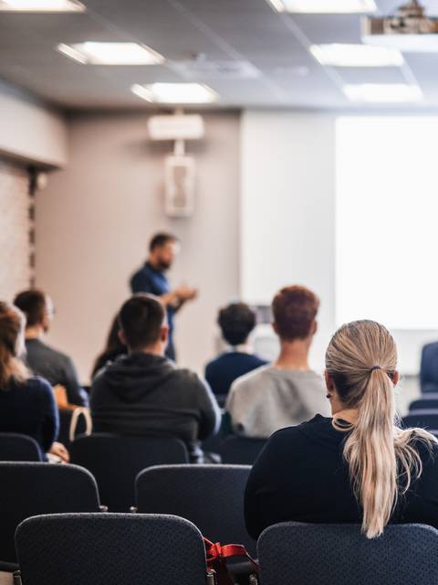 Un grupo de personas sentadas en una sala con un presentador delante de una pantalla blanca.