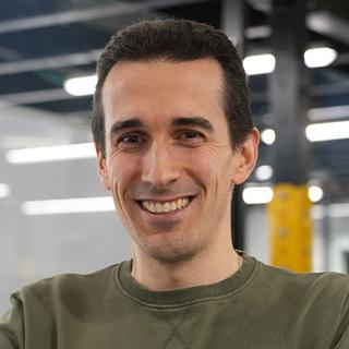 Un hombre sonriente viste un suéter de color verde en un entorno moderno.