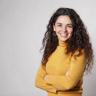 Una mujer sonriente con cabello rizado y una prenda amarilla posa con los brazos cruzados.