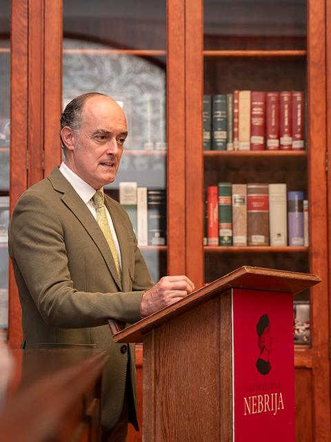Un hombre de pie frente a un podio en una biblioteca con estanterías llenas de libros.