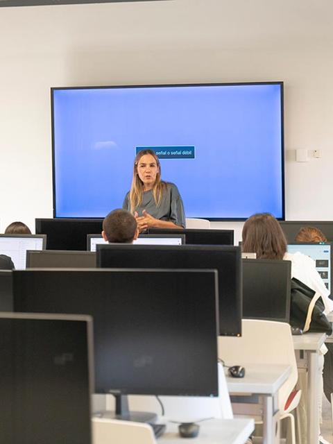 Una mujer está dando una charla frente a un grupo de estudiantes en un aula con computadoras.