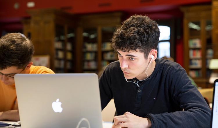 Un joven estudia concentrado frente a una computadora en una biblioteca.