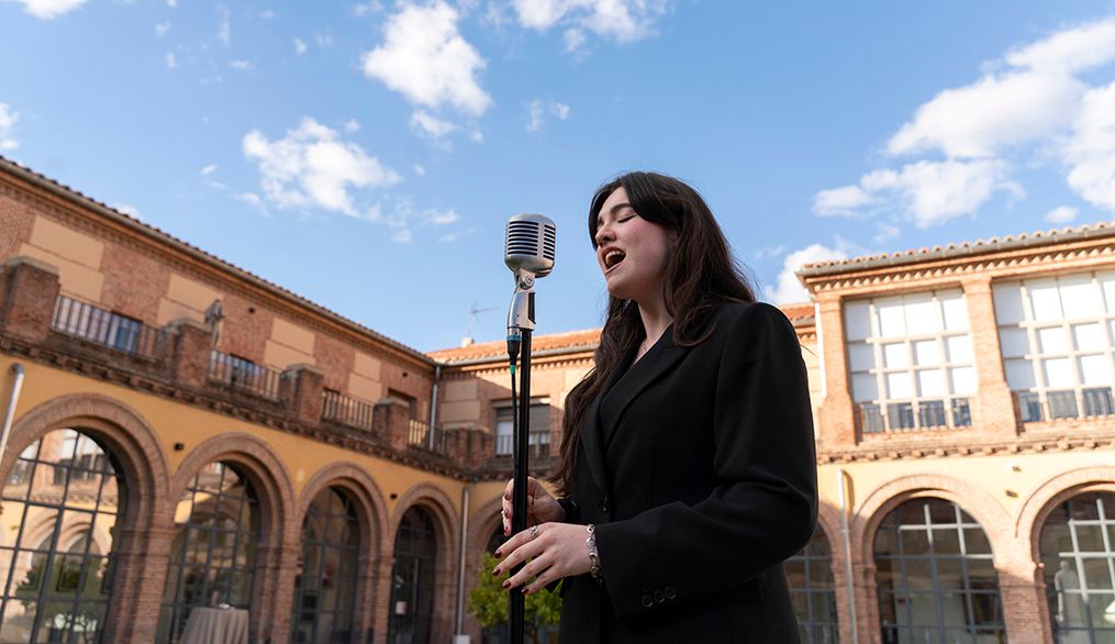 Una joven canta frente a un micrófono en un patio con arquitectura histórica bajo un cielo azul.