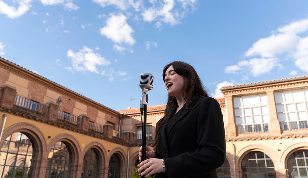 Una joven canta frente a un micrófono en un patio con arquitectura histórica bajo un cielo azul.