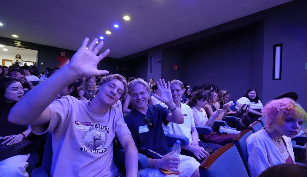 Un grupo de jóvenes sonríen y saludan desde sus asientos en un evento.