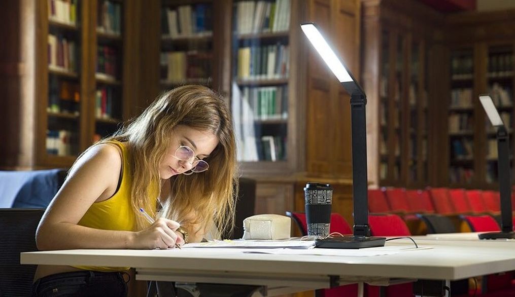 Una estudiante está escribiendo en una mesa iluminada en una biblioteca.