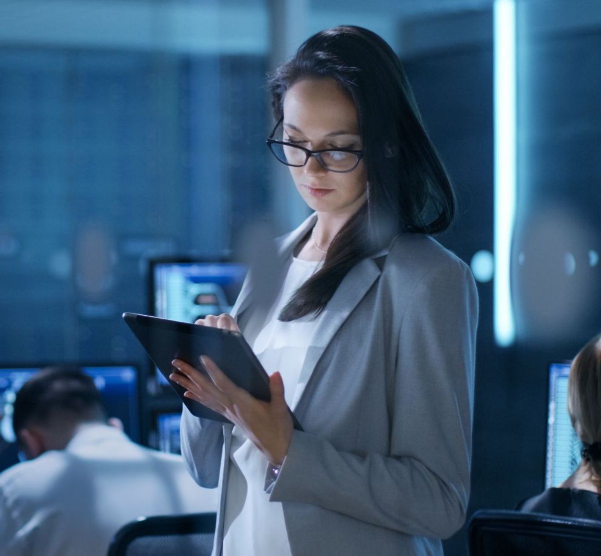 Una mujer con gafas sostiene una tableta y está concentrada, mientras en el fondo se pueden ver otras personas trabajando en computadoras.