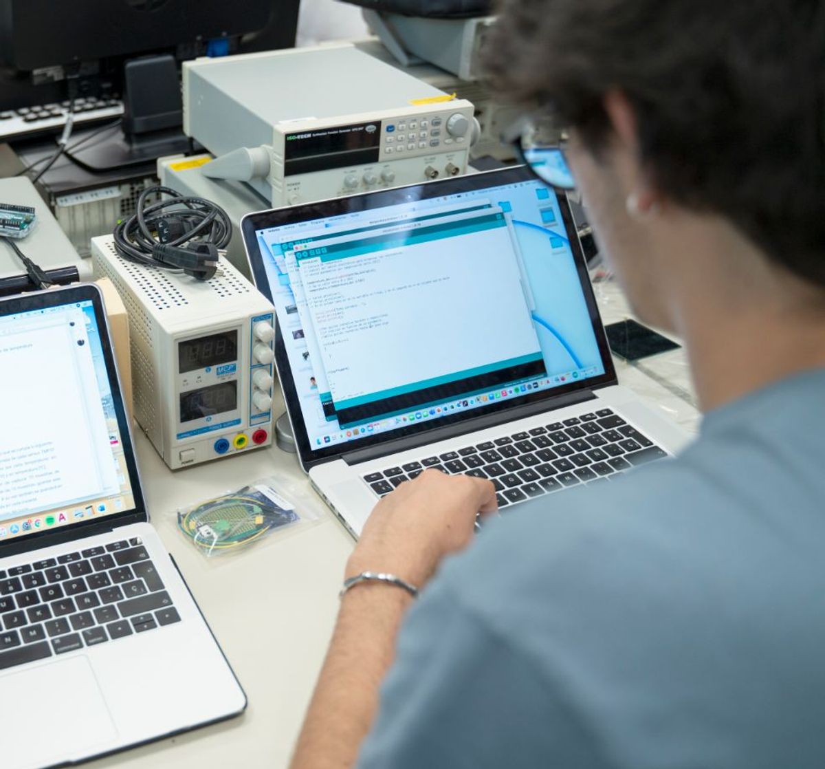Una persona trabaja en dos computadoras portátiles mientras está rodeada de equipos de laboratorio y componentes electrónicos.
