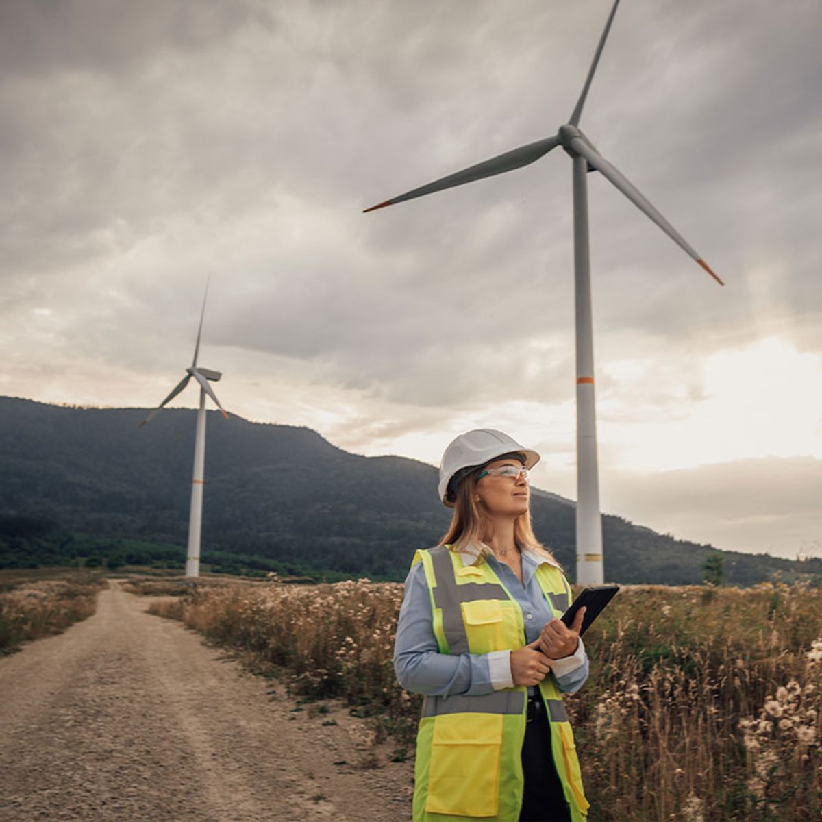 La imagen muestra a una mujer con casco y chaleco reflectante de pie en un camino rural, rodeada de turbinas eólicas bajo un cielo nublado.